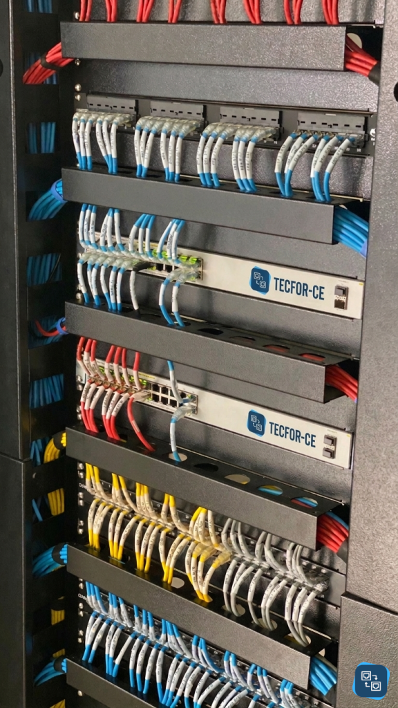 Visão em close de um rack de servidores da TECFOR-CE com organização impecável de cabos de rede coloridos e switches identificados em Fortaleza, Ceará. Infraestrutura profissional de TI.