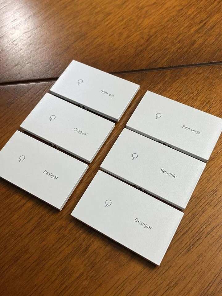 Seis teclas brancas de keypad para automação residencial da TECFOR-CE dispostas sobre uma mesa de madeira. Cada tecla possui um ícone de lâmpada e palavras gravadas a laser: Bom dia, Cheguei, Desligar, Bem vindo e Reunião