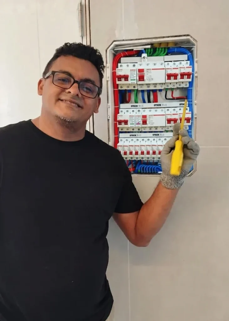 Um eletricista profissional com luva e chave de fenda posando ao lado de um quadro elétrico residencial moderno com fiação colorida e disjuntores.